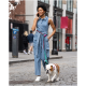 Eine Frau in einem Jeans-Overall führt einen Bulldoggen an der Leine entlang einer Kopfsteinpflasterstraße und spricht dabei am Telefon.