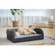 Fluffy tan-and-white dog lying on a gray bolster dog bed with small leather tag in a bright living room.
