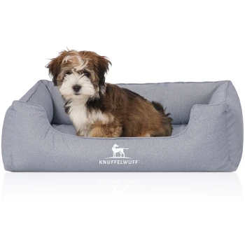 Ein kleiner, flauschiger Hund sitzt bequem in einem grauen Haustierbett mit dem Logo "KNUFFELWUFF" sichtbar auf der Vorderseite.