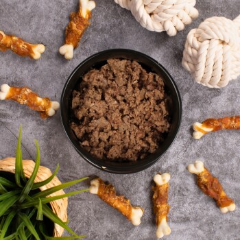 Eine schwarze Schüssel, gefüllt mit feuchtem Haustierfutter, steht auf einer grauen Oberfläche, umgeben von Hundekauartikeln und zwei Seilspielzeugen, mit grünen Pflanzenblättern in der Nähe.