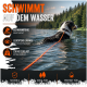 Nasser Hund steht im flachen Flusswasser mit dem Rücken zur Kamera, an eine orangefarbene Leine gebunden, mit Bäumen und Grün im Hintergrund.