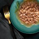 Une assiette turquoise contient de la nourriture humide pour animaux avec des morceaux de viande dans une sauce, garnie de romarin, à côté d'une fourchette dorée sur un tissu sombre.