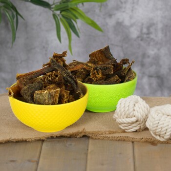 Eine schwarze Schüssel, gefüllt mit verschiedenen getrockneten Hundeleckereien, einschließlich Kaustangen und Sticks, umgeben von grünem Gras und zwei weißen Seilspielzeugen auf einem grauen Hintergrund.