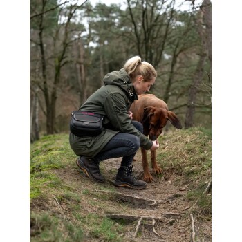 Eine Person in einer grünen Jacke kniet auf einem Schotterweg und füttert einen braunen Hund. Umgebende Bäume und Gras schaffen eine natürliche Umgebung.