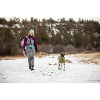 Eine Frau in einem lila Langarmshirt und grauen Hosen geht mit einem glücklichen Hund in einem grünen Mantel auf einem verschneiten Weg.