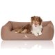 Ein glücklicher braun-weißer Hund liegt auf einem gemütlichen, braunen Hundebett mit dem Logo "KNUFFELWUFF" sichtbar auf der Vorderseite.