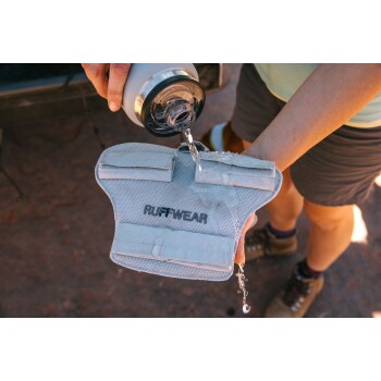 Eine Person gießt Wasser aus einer Flasche auf ein graues Netz-Hundegeschirr mit der Aufschrift 'RUFFWEAR', während Wasser vom Geschirr tropft.