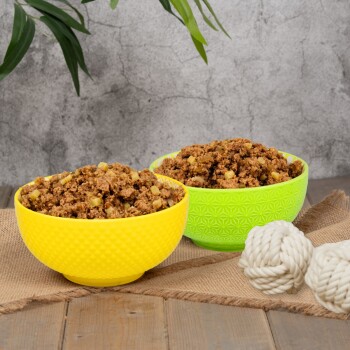Zwei Schalen mit stückigem Nassfutter für Hunde mit Mais, serviert in gelber und grüner strukturierter Schale, auf einer Jute-Matte mit zwei weißen Tau-Spielzeugen