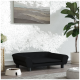 Ein stilvolles schwarzes Haustiersofa mit abgerundeten Armen und Knöpfen, das auf einem Holzboden neben einer weißen Futterschüssel und einer Topfpflanze steht.