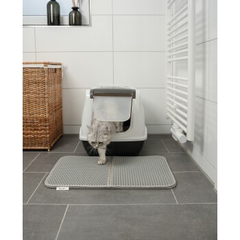 Eine graue Katze, die aus einer modernen Katzentoilette auf eine strukturierte graue Matte in einem sauberen, gefliesten Badezimmer mit einem geflochtenen Korb in der Nähe tritt.