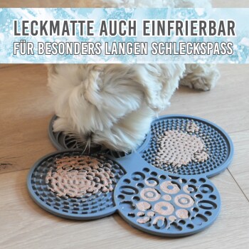 Ein kleiner weißer Hund leckt eine graue Silikonmatte mit verschiedenen Texturen und dem Text "Leckmatte auch einfrierbar für besonders langen Schleckspass."