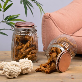 Ein Glasbehälter mit Hundeleckerlis auf einem Holztisch, mit verstreuten Leckerlis und einem gewickelten Seilspielzeug in der Nähe, vor einem rosa Sofa.