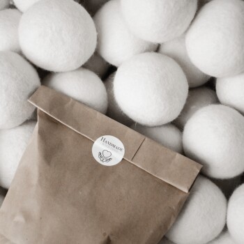 Ein Paket handgefertigter weißer Wolltrocknerbälle liegt auf einem Haufen ähnlicher Bälle, mit einem Label, das "HANDMADE WITH LOVE" liest.