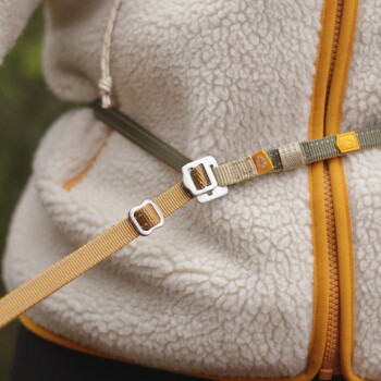 Nahaufnahme gekreuzter olivgrüner und senffarbener verstellbarer Hundegeschirrriemen mit Metallverschlüssen und Pfotenanhänger auf einer cremefarbenen Sherpa-Jacke.
