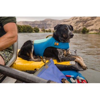 Ein Hund, der eine blaue Schwimmweste trägt, ruht auf einem gelben Schwimmgerät neben einem Fluss, mit einer malerischen Kulisse von Hügeln.