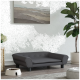 Ein stilvolles graues Haustiersofa mit Knopfdetails steht auf einem Holzboden, neben einer weißen Futterschüssel, die mit Trockenfutter gefüllt ist, und einer Topfpflanze.