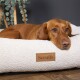 Ein entspannter brauner Hund ruht seinen Kopf auf einem plüschigen, cremefarbenen Hundebett mit einem braunen "Scruffs"-Etikett, vor einem dunklen Hintergrund.
