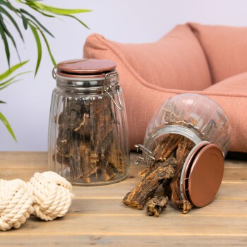 Zwei Glasbehälter mit Hundeleckerlis, einer umgekippt, neben einem Seilspielzeug auf einem Holztisch mit einer rosa Couch im Hintergrund.