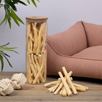 Ein Glasbehälter, gefüllt mit Hundekauartikeln, steht neben einem plüschigen, rosa Haustierbett, mit zusätzlichen Kauartikeln, die auf einem Holztisch verstreut sind.