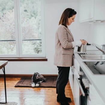 Eine Frau in einem karierten Blazer steht neben einem Küchenspülbecken und gießt Wasser aus einem Wasserkocher, während ein Hund auf einer Matte in der Nähe ruht.