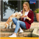 Une femme en cardigan bordeaux est assise sur des marches en pierre, tenant un paquet de friandises pour chiens Vitakraft tandis qu'un golden retriever s'approche d'elle avec empressement.