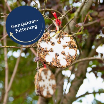 Ein schneeflockenförmiger Vogelfutterornament auf einem Holzbrett, mit einem roten Band und einem goldenen Abzeichen mit der Aufschrift 'Beste Qualität.'