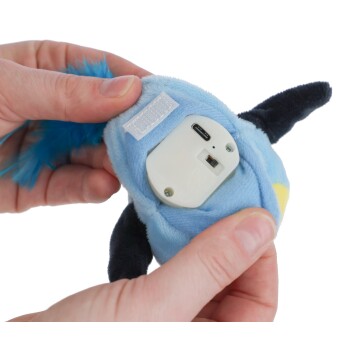 Hände halten ein blaues Plüschvogelspielzeug mit herausnehmbarem elektronischem Modul, das Ladeanschluss und Schalter zeigt.