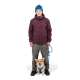 Eine Person in einer kastanienbraunen Jacke und blauen Mütze steht mit einem an der Leine geführten Corgi zu ihren Füßen, vor einem weißen Hintergrund.