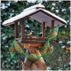 Vogelhaus aus Holz mit Schnee auf dem Dach, zwei kleine Vögel füttern und hängende grüne Netzbeutel gefüllt mit Samen.
