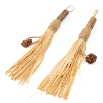 Zwei natürliche Kauspielzeuge für Haustiere aus Stroh und Holz, mit ausgefransten Enden und einem kleinen Ball an der Spitze, mit Schnur gebunden.