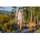 Eine Frau in einem grauen Tanktop und grünen Shorts führt einen braunen Hund an der Leine auf einem bewaldeten Weg, umgeben von Bäumen und Sträuchern.
