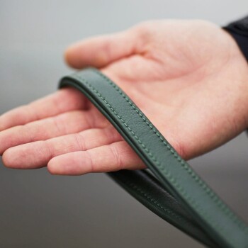Eine Hand, die eine grüne Lederleine für Hunde mit sichtbaren Nähdetails hält, vor einem verschwommenen Hintergrund.