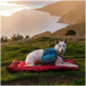 Ein weißer Hund mit einem blauen Rucksack ruht auf einer roten Decke und blickt auf eine malerische Küstenlandschaft bei Sonnenuntergang.