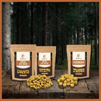 Drei Tüten Wildfang-Käse-Snacks für Hunde, beschriftet mit "Harties", "Softies" und "Käsebrocken", liegen auf einer Holzoberfläche in einem dunklen Wald.