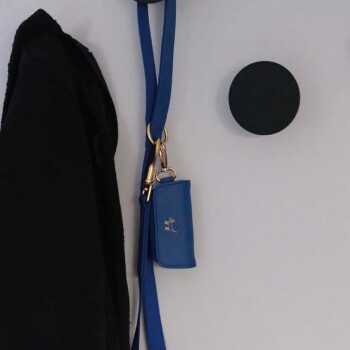 Eine blaue Hundeleine hängt an einer Wand neben einer schwarzen Jacke, mit einem kleinen blauen Beutel, der eine goldene Hundesilhouette zeigt.