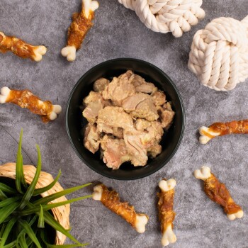 Eine schwarze Schüssel, gefüllt mit Stückchen Haustierfutter, steht auf einer grauen Oberfläche, umgeben von Hundekau-Knochen und einer kleinen grünen Pflanze.