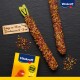 Zwei bunte Vogelleckerlis, die mit Samen und Körnern überzogen sind, etwa 15 cm lang und 2-3 cm im Durchmesser, neben einer gelben Vitakraft-Box.