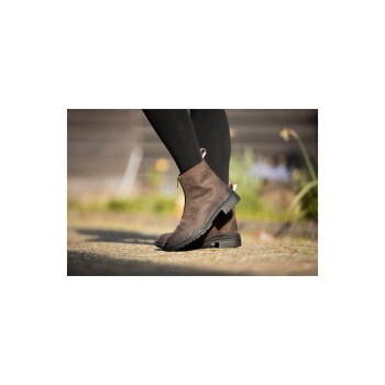 Eine Nahaufnahme von den Beinen einer Person, die braune Knöchelstiefel und schwarze Leggings trägt, auf einem Steinweg mit Grün im Hintergrund.