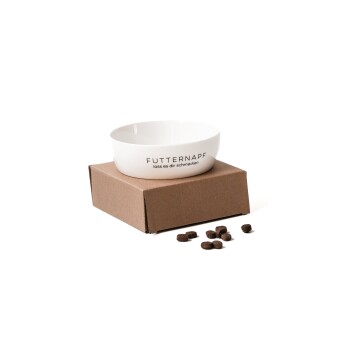 Eine weiße Futterschüssel für Haustiere, beschriftet mit "FUTTERNAPF lass es dir schmecken", steht auf einem braunen Karton, mit verstreuten Futterstücken in der Nähe.
