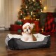 Ein kleiner Hund in einem Weihnachtsmannhut sitzt in einem grauen Haustierbett, umgeben von einem geschmückten Weihnachtsbaum und bunten Geschenken.