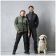 Ein Mann und eine Frau stehen nebeneinander, beide tragen stilvolle Outdoor-Jacken und Hosen, während ein Golden Retriever neben ihnen sitzt.