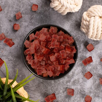 Eine schwarze Schüssel gefüllt mit kleinen, gewürfelten roten Haustierleckereien, umgeben von verstreuten Stücken und zwei geknoteten Seilspielzeugen auf einer grauen Oberfläche.