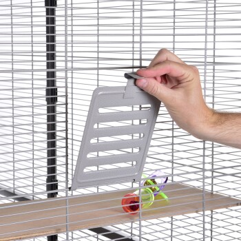 Eine Hand hält ein graues Schiebetürpaneel für einen Haustierkäfig, mit bunten Spielzeugen, die im Käfig auf einem Holzregal sichtbar sind.