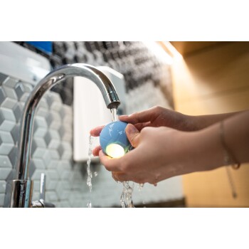 Eine Person wäscht ein blaues Haustierspielzeug unter fließendem Wasser aus einem Wasserhahn, während Wasser um ihre Hände spritzt.