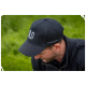 Eine Person trägt eine schwarze Kappe mit einem weißen "U"-Logo, sitzt in einem grasbewachsenen Bereich und konzentriert sich auf eine unsichtbare Aufgabe.