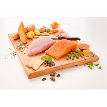 Ein Holzschneidebrett hält frische Zutaten für Tierfutter: Lachs, Huhn, Süßkartoffel, Erbsen, Apfel, Ingwer, Honig und Kräuter.
