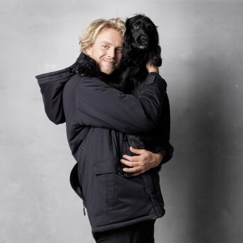 Eine Person in einer schwarzen Jacke hält einen schwarzen Hund nah, was eine warme Bindung zwischen ihnen vor einem grauen Hintergrund zeigt.