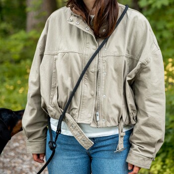 Person in beiger Jacke und Jeans trägt eine schwarze Seilleine quer über der Brust; Schnauze eines Hundes links sichtbar.