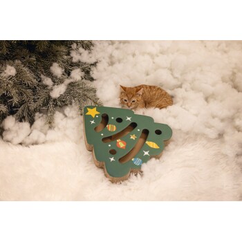 Orange tabby cat lying on fluffy white bedding next to a green Christmas tree-shaped cat scratcher with ornaments.