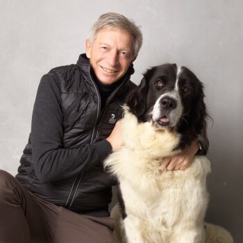 Eine Person in einer schwarzen Weste sitzt neben einem großen, flauschigen Hund mit einem schwarz-weißen Fell, beide schauen in die Kamera.